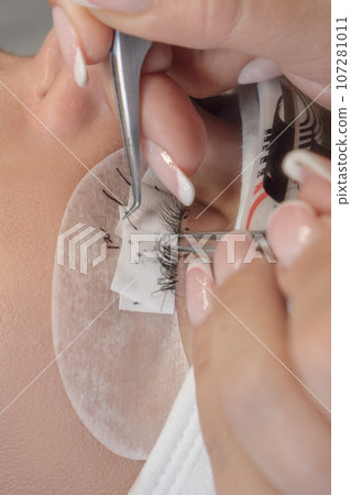 Close up of lashmaker with tweezers parting cilia for sticking artificial eyelash on eyelid of client with closed eyes. 107281011