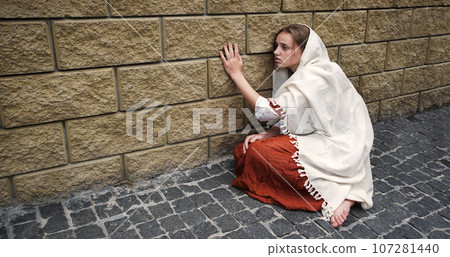 A girl in poor clothes sits on the ground 107281440