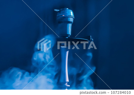 Hookah head, clay bowl and pipe, smoking object in smoke and blue light, relaxation atmosphere Hookah head, clay bowl and pipe, smoking object in smoke and blue light, relaxation atmosphere 107284010
