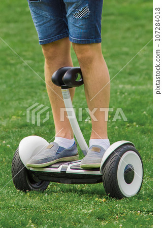 The legs of an adult man stand on a hoverboard against the background of grass in the park The legs of an adult man stand on a hoverboard against the background of grass in the park 107284018