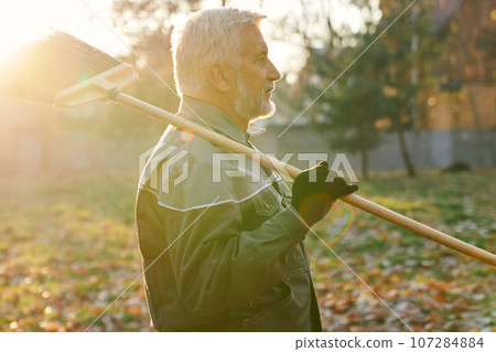 Senior maintenance worker looking away, while keeping broom on shoulder in warm autumn day. Side view of bearded man in age walking with broom, with sunlit background. Maintenance work, fall concept. Senior maintenance worker looking away, while keeping broom on shoulder in warm autumn day. Side view of bearded man in age walking with broom, with sunlit background. Maintenance work, fall concept. 107284884