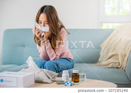 Sick woman. Asian young girl cold sick she sneeze with tissue paper on sofa, beautiful female health problem blowing nose use pharmacy kit box delivery service from hospital, delivery pharmacy concept Sick woman. Asian young girl cold sick she sneeze with tissue paper on sofa, beautiful female health problem blowing nose use pharmacy kit box delivery service from hospital, delivery pharmacy concept 107285240
