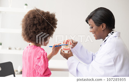 African American dentist explaining to mixed-race afro child how to brush teeth. The doctor and healthcare concept for children. African American dentist explaining to mixed-race afro child how to brush teeth. The doctor and healthcare concept for children. 107285935