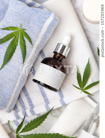 Dropper bottle with blank label near green cannabis leaves top view, CBD cosmetic mockup 107285969