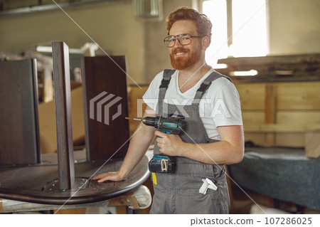 Smiling carpenter fixing table with electric drill standing in carpentry workshop and looks camera 107286025
