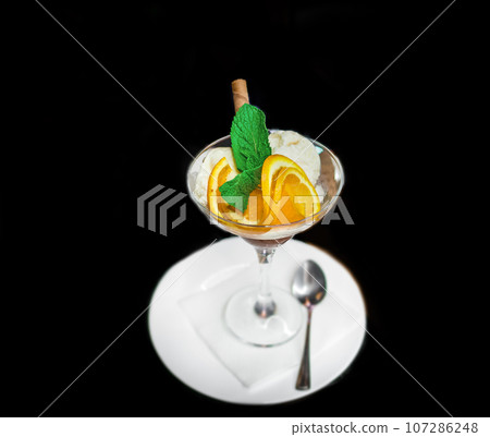 Beautiful serving of scoops of ice cream with slices of orange and mint, ice cream in a glass and saucer with a spoon on a black background 107286248
