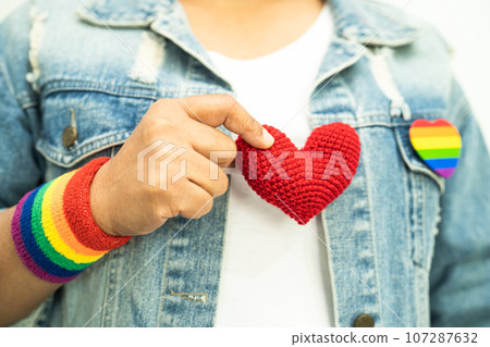 Asian lady wearing rainbow flag wristbands and hold red heart, symbol of LGBT pride month celebrate annual in June social of gay, lesbian, bisexual, transgender, human rights. Asian lady wearing rainbow flag wristbands and hold red heart, symbol of LGBT pride month celebrate annual in June social of gay, lesbian, bisexual, transgender, human rights. 107287632