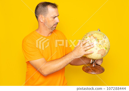 Hispanic man with beard in his 40s searching for a location on a world globe, isolated on yellow background. 107288444