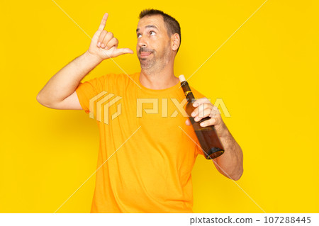 Hispanic man with beard in his 40s making a go drunk gesture with a bottle of wine in his hand, isolated on yellow studio background. 107288445