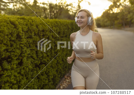Smiling blonde woman wearing modern wireless headphones running through the park on a bright sunny summer day. A happy girl with a sweet smile runs and listens to good music. Lifestyle. Smiling blonde woman wearing modern wireless headphones running through the park on a bright sunny summer day. A happy girl with a sweet smile runs and listens to good music. Lifestyle. 107288517