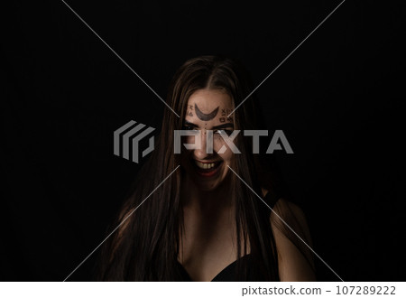 Evil laughing at the top of her voice, the witch looks into the camera on a black background in the twilight. On Halloween, a female witch grinning in a terrible grin looks at the camera. Evil laughing at the top of her voice, the witch looks into the camera on a black background in the twilight. On Halloween, a female witch grinning in a terrible grin looks at the camera. 107289222