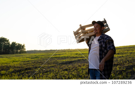 The farmer looks in surprise towards the empty space for insertion while standing at sunset in a field with a box of potatoes. The concept of agriculture, harvesting. A man rejoices in the harvest. The farmer looks in surprise towards the empty space for insertion while standing at sunset in a field with a box of potatoes. The concept of agriculture, harvesting. A man rejoices in the harvest. 107289236