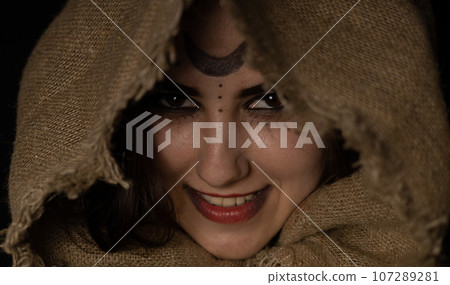 A grinning witch peeks out from under the hood at Halloween night. A beautiful female witch with a hood thrown over her head smirks while looking at the vamer on a black background. Close-up. 107289281