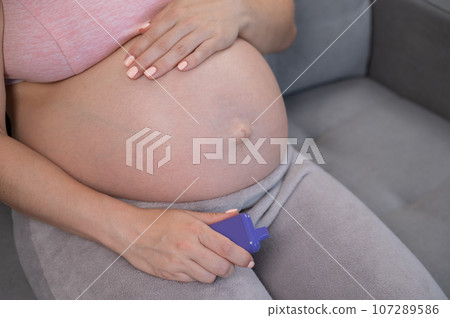 A pregnant woman smokes a vape while sitting on the couch. Close-up of the abdomen. 107289586