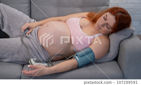A pregnant woman measures blood pressure with a tonometer while lying on the couch. Hypertension.  107289591