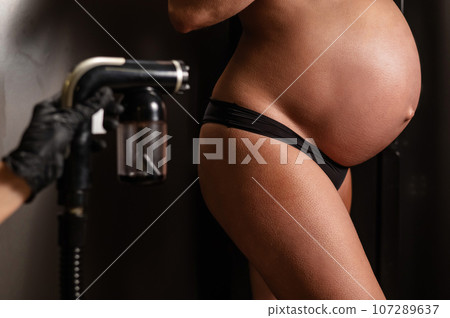 Pregnant woman getting an instant tan. The master applies bronzer with a spray. 107289637