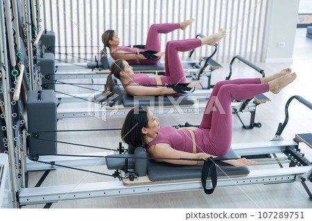 Three Asian women in pink sportswear doing pilates exercises with a reformer bed.  107289751