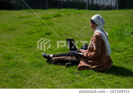 A young woman wearing a hijab sits on a lawn and uses a laptop outdoors. A young woman wearing a hijab sits on a lawn and uses a laptop outdoors. 107289752