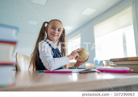 Caucasian schoolgirl sits at her desk at school and studies the globe. Caucasian schoolgirl sits at her desk at school and studies the globe. 107290063