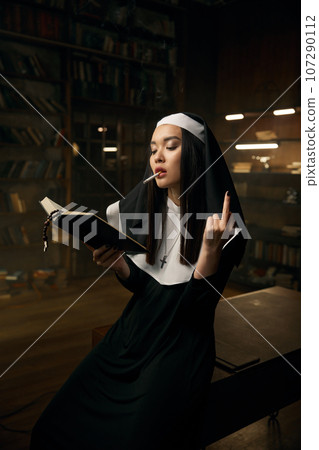 Nun woman reading bible, smoking cigarette and showing obscene hand gesture 107290112