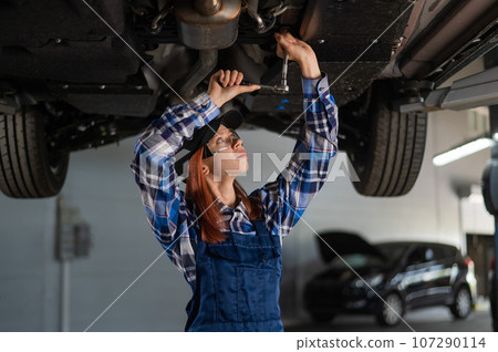 Female mechanic unscrew the nuts on the bottom of the car that is on the lift. A girl at a man's work. Female mechanic unscrew the nuts on the bottom of the car that is on the lift. A girl at a man's work. 107290114