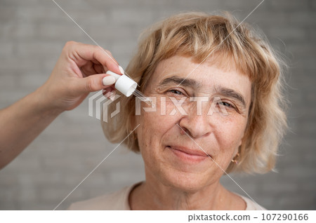 Close-up portrait of an old woman applying hyaluronic acid serum with a pipette. Anti-aging face care. Close-up portrait of an old woman applying hyaluronic acid serum with a pipette. Anti-aging face care. 107290166