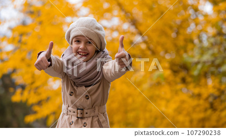 Happy caucasian girl in a beige coat and beret walks in the park in autumn. Schoolgirl showing thumbs up. Happy caucasian girl in a beige coat and beret walks in the park in autumn. Schoolgirl showing thumbs up. 107290238