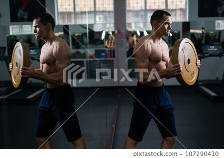Muscular shirtless man doing exercise with weight plate in gym. Muscular shirtless man doing exercise with weight plate in gym. 107290410