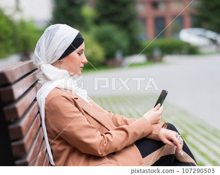 Young woman in hijab uses a smartphone while sitting on a bench outdoors. Young woman in hijab uses a smartphone while sitting on a bench outdoors. 107290503