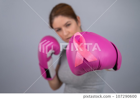 Caucasian woman in pink boxing gloves with a pink ribbon on her chest on a gray background. Fight against breast cancer. Caucasian woman in pink boxing gloves with a pink ribbon on her chest on a gray background. Fight against breast cancer. 107290600
