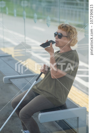 Elderly blind woman sitting at a bus stop and using a smartphone.  Elderly blind woman sitting at a bus stop and using a smartphone.  107290615