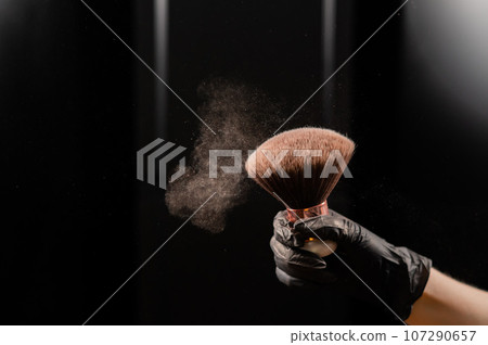 A woman shakes a large powder brush with clouds of dust on a black background. Treatment after instant tanning A woman shakes a large powder brush with clouds of dust on a black background. Treatment after instant tanning 107290657