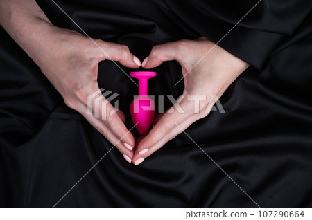 Woman making a heart shape with her palms around a pink butt plug on a black silk sheet. Woman making a heart shape with her palms around a pink butt plug on a black silk sheet. 107290664