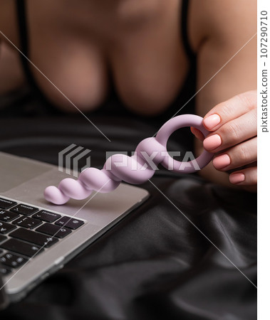 Woman holding lilac anal beads next to laptop while lying on black sheet. Woman holding lilac anal beads next to laptop while lying on black sheet. 107290710