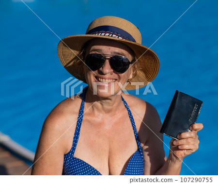 An elderly woman in a hat and sunglasses holds a passport while sitting by the pool. Vacation in retirement.  107290755