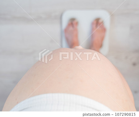 Top view of a pregnant woman with a bare tummy standing on an electronic scale. Top view of a pregnant woman with a bare tummy standing on an electronic scale. 107290815