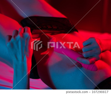 A faceless woman lies in a bedroom in a blue-red neon light and uses a pink vibrator. Close-up of the abdomen. A faceless woman lies in a bedroom in a blue-red neon light and uses a pink vibrator. Close-up of the abdomen. 107290817
