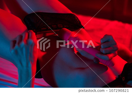 A faceless woman lies in a bedroom in a blue-red neon light and uses a pink vibrator. Close-up of the abdomen. A faceless woman lies in a bedroom in a blue-red neon light and uses a pink vibrator. Close-up of the abdomen. 107290821