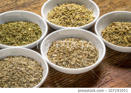 herb spices - dried leaves of parsley, rosemary, thyme, oregano and basil, collection of small ceramic bowls on a rustic wood 107291051