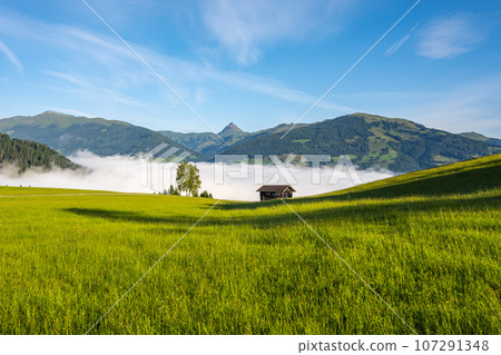 Alpine mountain landscape with green meadows, rural hayloft and Grosser Rettenstein Mountain on background. Sunny morning with weather inversion in the valley. Kitzbueheler Alps, Austria 107291348