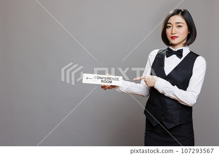 Asian woman receptionist holding conference room directional tabletop signage and pointing with finger portrait. Hotel employee in uniform showing boardroom steel sign and looking at camera Asian woman receptionist holding conference room directional tabletop signage and pointing with finger portrait. Hotel employee in uniform showing boardroom steel sign and looking at camera 107293567