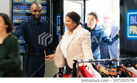 Guard open doors on black friday event, allowing access to clothes in shopping center. Diverse people rushing inside to buy items at half price, findind best discounts on clearance. 107293574