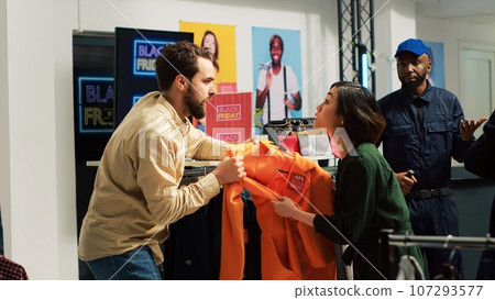 Black Friday fight in clothing store. Mad shoppers fighting over clothes during seasonal sales at mall, angry customers pulling clothing item and arguing, various brands on discount. 107293577