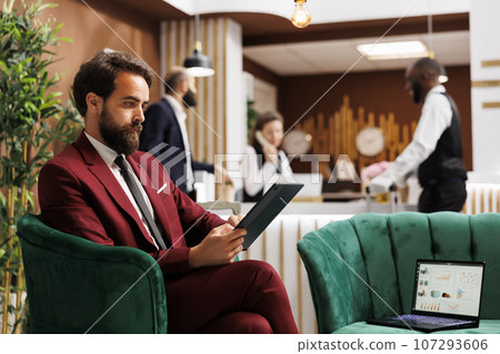 Employee looks over meeting notes waiting to do check in before attending important international conference. Young businessman at hotel travelling for work, reading documents in lobby. Employee looks over meeting notes waiting to do check in before attending important international conference. Young businessman at hotel travelling for work, reading documents in lobby. 107293606