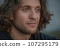 Close-up of young adult handsome man with long curly hair and stubble looks forward outdoor 4K 107295179