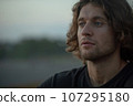 Close-up of young adult handsome man with long curly hair and stubble looks forward outdoor 4K 107295180