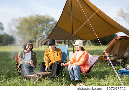 Young men and women enjoying camping, bonfire, three people laughing while roasting marshmallows, outdoor image Young men and women enjoying camping, bonfire, three people laughing while roasting marshmallows, outdoor image 107297712