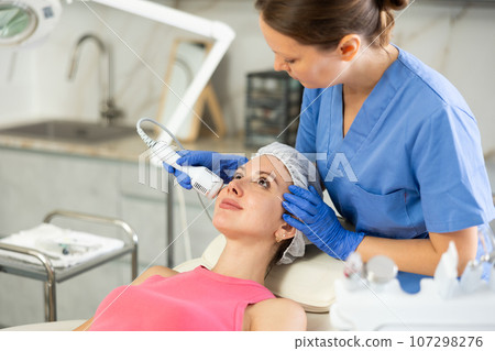 Adult woman cosmetologist doing whitening procedure to young woman Adult woman cosmetologist doing whitening procedure to young woman 107298276