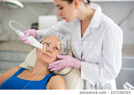 Young beautician doing procedure for face lifting and Soften skin keratin, deep clear skin dirt of adult woman laying in salon. Hardware cosmetology Young beautician doing procedure for face lifting and Soften skin keratin, deep clear skin dirt of adult woman laying in salon. Hardware cosmetology 107298565