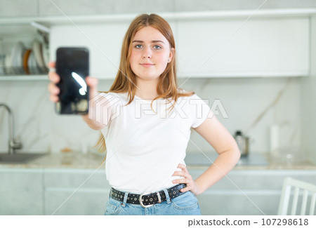 Smiling teenage girl presenting smartphone with blank black screen with copy space in modern cuisine. Free copy space for your design or advertisement Smiling teenage girl presenting smartphone with blank black screen with copy space in modern cuisine. Free copy space for your design or advertisement 107298618